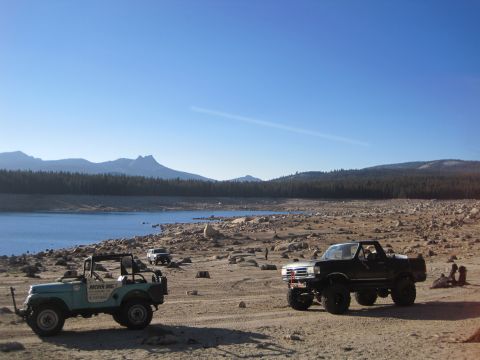 1989 Ford Bronco - Two off-road vehicles are parked near a lake with mountains in the distance under a clear blue sky.
