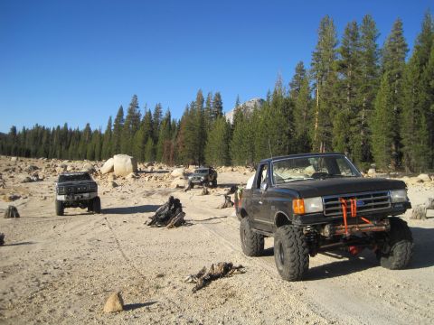 1989 Ford Bronco - Several off-road vehicles are parked on a dirt track in a rocky, wooded area under a clear blue sky.