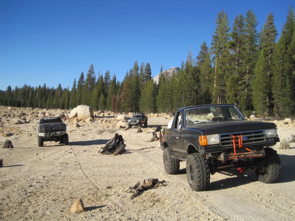 Black Ford F-Series pickup truck with lifted suspension and off-road tires parked on a dirt road with trees and rocks in the background. Two other SUVs are visible further back