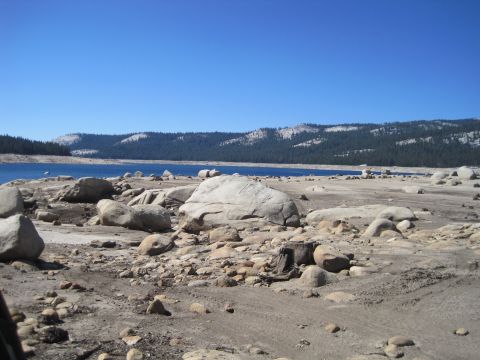 1989 Ford Bronco - A dry, rocky shoreline borders a calm blue lake, with forested hills in the background under a bright, cloudless sky.
