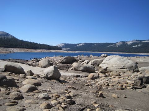 1989 Ford Bronco - A wide view of a rocky shoreline leading to a blue lake, with forested hills in the background under a bright blue sky.