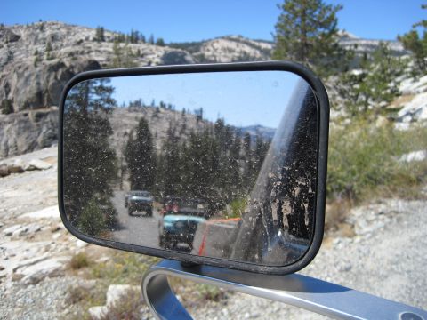 1989 Ford Bronco - A reflection in a dusty side mirror shows multiple vehicles driving on a road through a mountainous, wooded area.