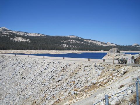 1989 Ford Bronco - A lone SUV is parked on the road atop a rocky dam, overlooking a vast blue lake under a clear sky.