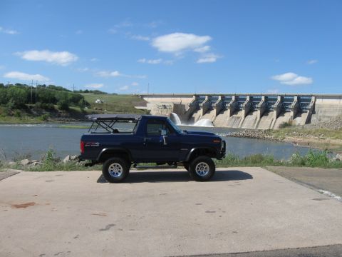 1982 Ford Bronco - A blue Ford Bronco is parked on a concrete pad with a large dam and reservoir in the background.