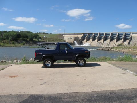 1982 Ford Bronco - A dark blue Ford Bronco is parked on a concrete ramp with a dam and water in the background.