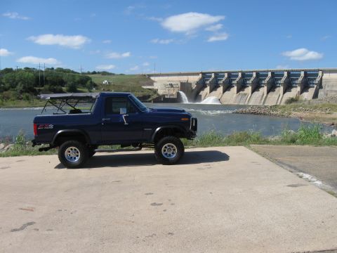 1982 Ford Bronco - A blue 4x4 SUV is parked on a concrete pad with a dam and water cascading in the background.