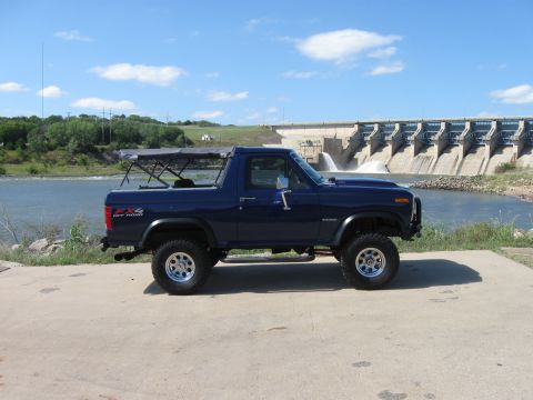 1982 Ford Bronco - A dark blue Ford Bronco is parked on a concrete pad in front of a large dam with water cascading down.