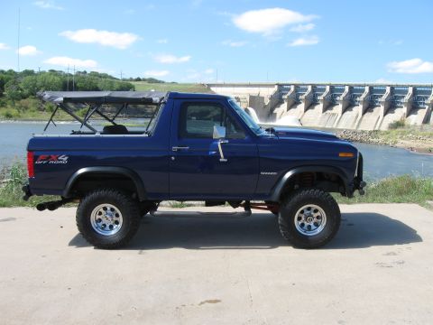 1982 Ford Bronco - This lifted blue Ford Bronco is parked in front of a large dam, with water flowing over the spillways.