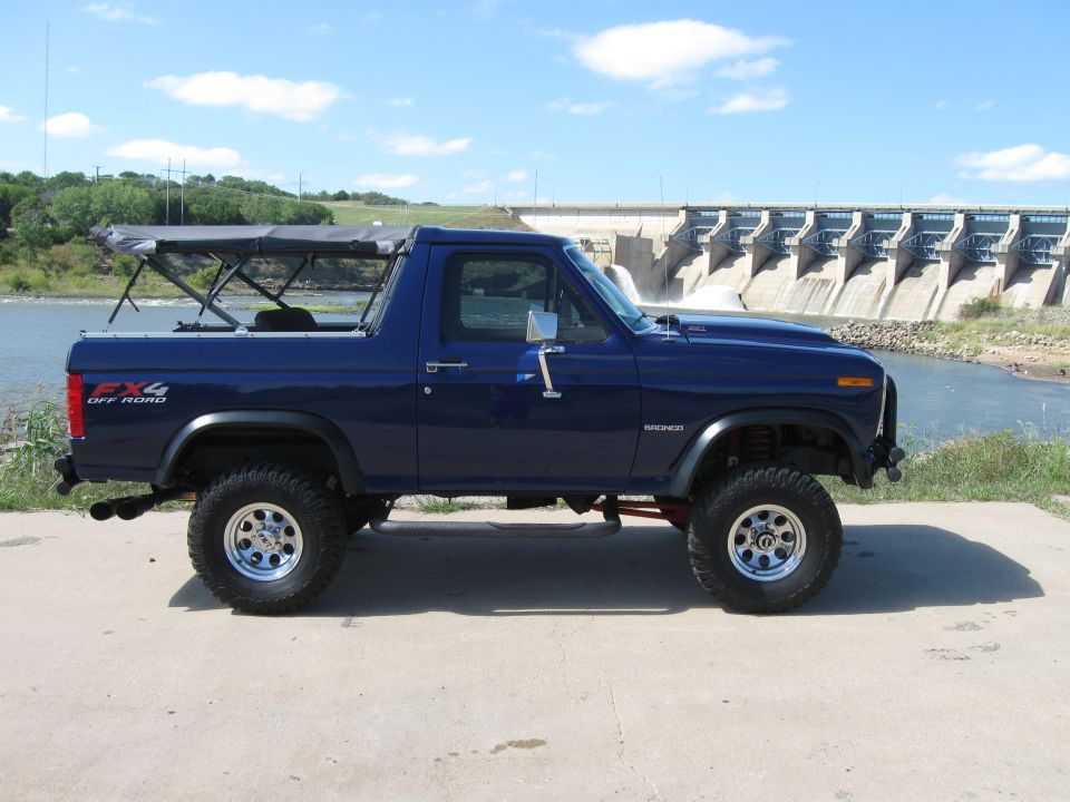 Blue Ford Bronco with a soft top, lifted suspension, and aftermarket wheels parked on concrete in front of a dam