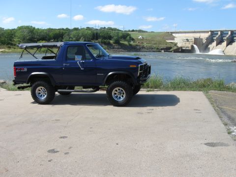 1982 Ford Bronco - This blue 1982 Ford Bronco features a removable soft top and is seen parked near a large dam with water flowing.