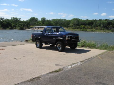 1982 Ford Bronco - A lifted blue Ford Bronco sits parked on a concrete pad next to a body of water under a clear blue sky.