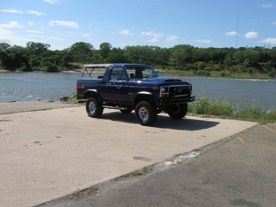 Blue Ford Bronco with aftermarket hood scoop and bumper parked on concrete near a river