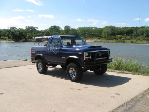 1982 Ford Bronco - A blue Ford Bronco is parked on a concrete surface next to a river under a clear sky.