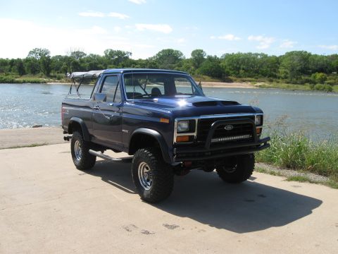 1982 Ford Bronco - This lifted 1982 Ford Bronco features a custom bumper and light bar, parked near the water on a sunny day.