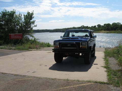1982 Ford Bronco - A blue Ford Bronco sits on a boat ramp with a lake in the background under a partly cloudy sky.