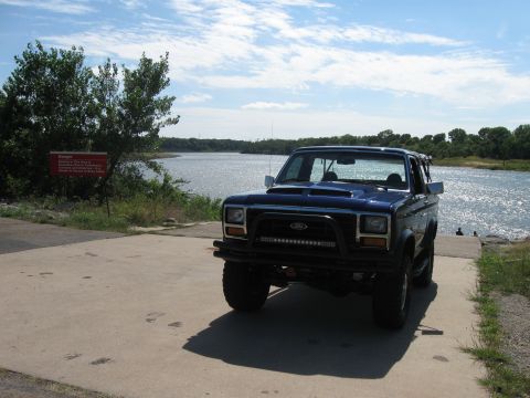 1982 Ford Bronco - A blue SUV is parked on a concrete ramp next to a body of water under a cloudy sky.