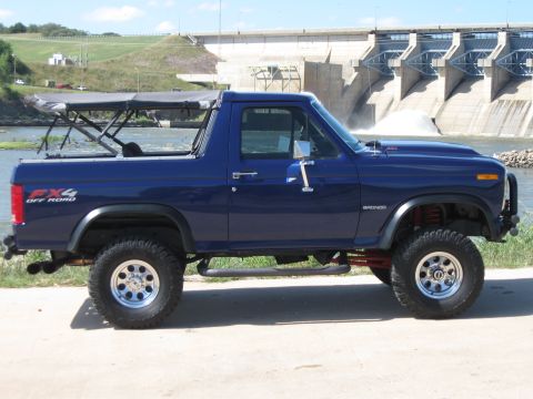 1982 Ford Bronco - Side view of a lifted 1982 Ford Bronco with a soft top, parked near a large dam.