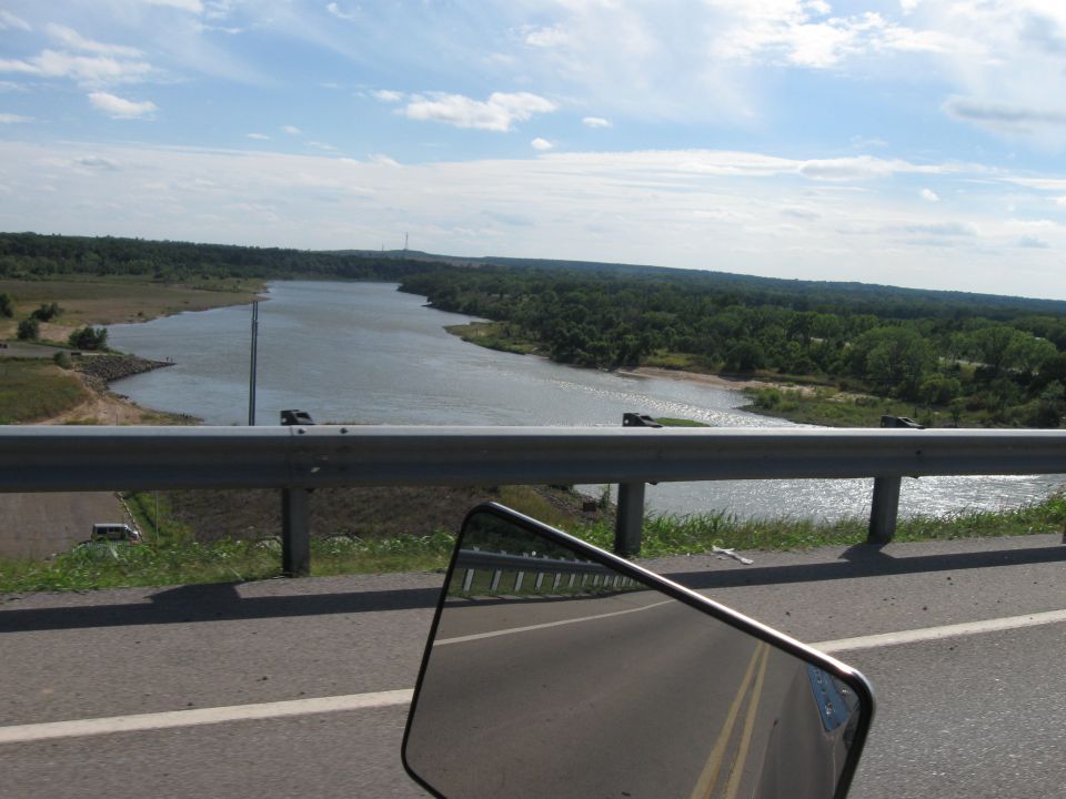 Side mirror reflection shows a road and a wide river with trees on the far bank