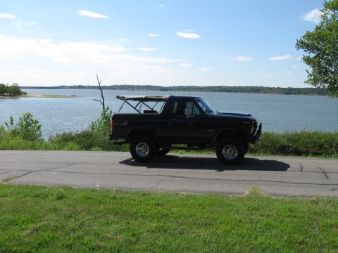 1982 Ford Bronco - A black Ford Bronco with a soft top is seen driving on a road beside a large body of water under a blue sky.