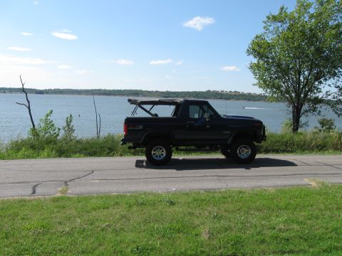 1982 Ford Bronco - A black 1982 Ford Bronco with its soft top partially deployed cruises down a road next to a large body of water.