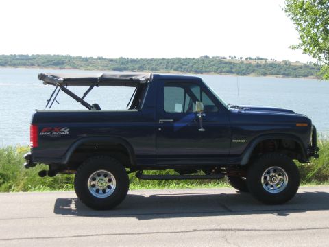 1982 Ford Bronco - Side view of a dark blue 1982 Ford Bronco with a soft top, parked near a body of water.