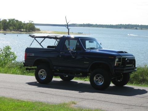 1982 Ford Bronco - This lifted Ford Bronco is parked on a paved road next to a large body of water with trees in the background.