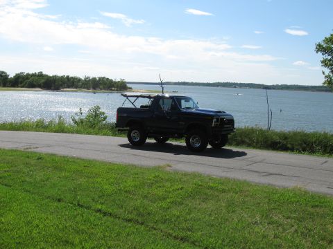 1982 Ford Bronco - A dark SUV with a roof rack drives along a road beside a large body of water under a partly cloudy sky.