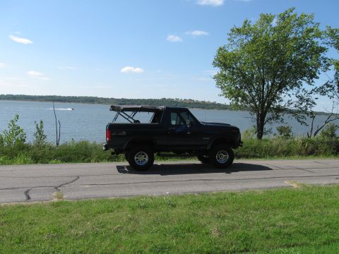 1982 Ford Bronco - Black 1982 Ford Bronco cruises down a road beside a large body of water under a clear sky.