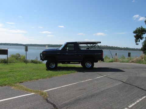 1982 Ford Bronco - A dark blue Ford Bronco is parked on the side of a road next to a body of water under a clear blue sky.
