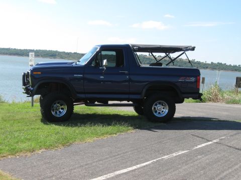 1982 Ford Bronco - A lifted, dark blue Ford Bronco with a soft top is parked by the water under a clear sky.