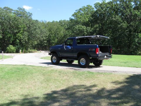 1982 Ford Bronco - Blue 1982 Ford Bronco with its soft top removed, parked on a dirt road with trees in the background.