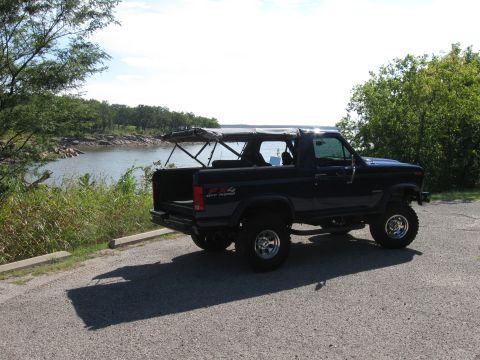 1982 Ford Bronco - A dark blue Ford Bronco is parked on a gravel lot next to a body of water under a partly cloudy sky.
