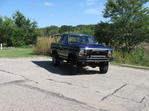 1982 Ford Bronco - This lifted Ford truck is parked on a paved surface with trees and water in the background.