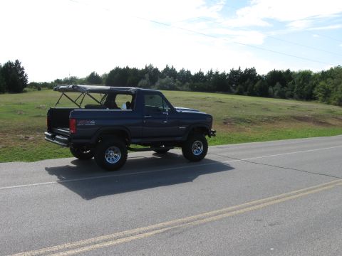 1982 Ford Bronco - A lifted blue Ford Bronco with its soft top partially open is seen driving on a paved road with a grassy hill in the background.