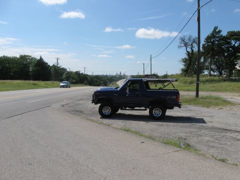 1982 Ford Bronco - A dark blue SUV sits parked on the shoulder of a highway under a bright blue sky.