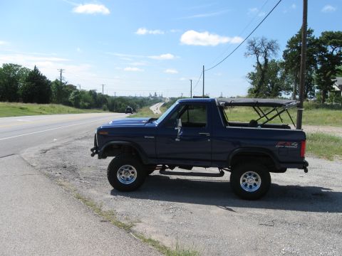 1982 Ford Bronco - A dark blue 1982 Ford Bronco with a soft top and '4x4 OFF ROAD' decal is parked on the side of a road.