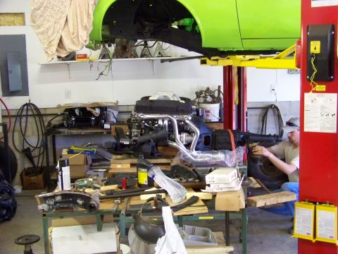1974 Dodge Challenger - A mechanic works on parts in a cluttered garage, with a car body suspended on a lift.