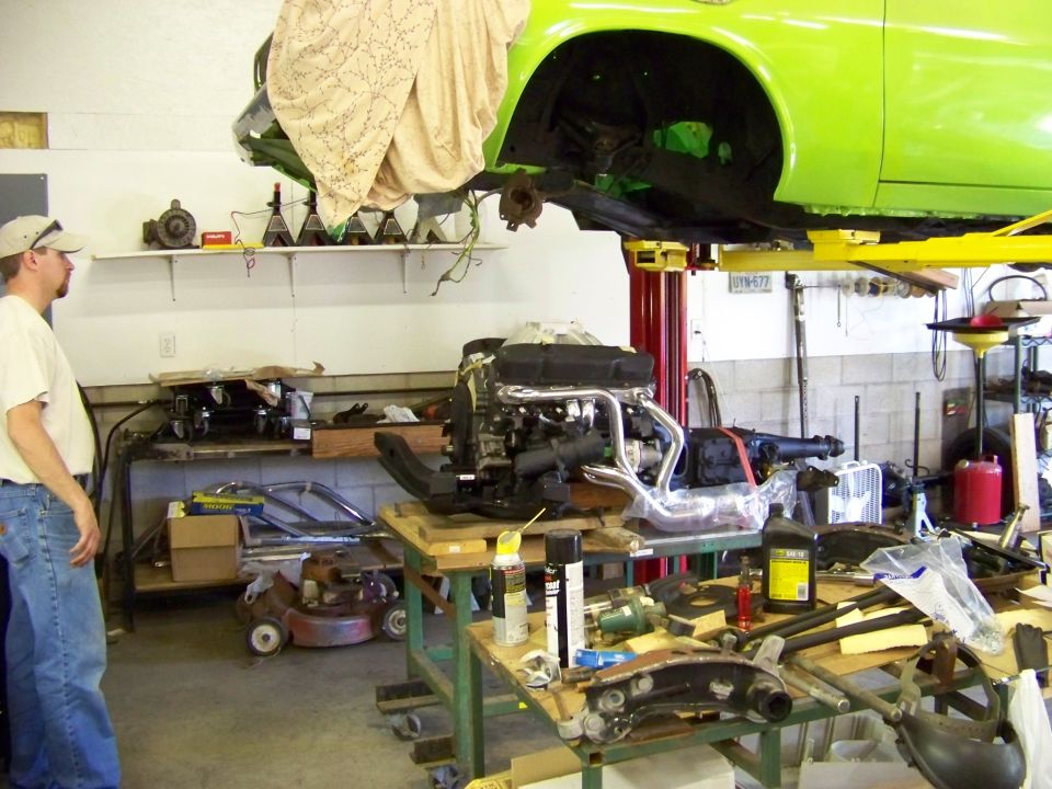 Engine on a stand with a green car body lifted above it in a garage. Parts and tools are scattered around