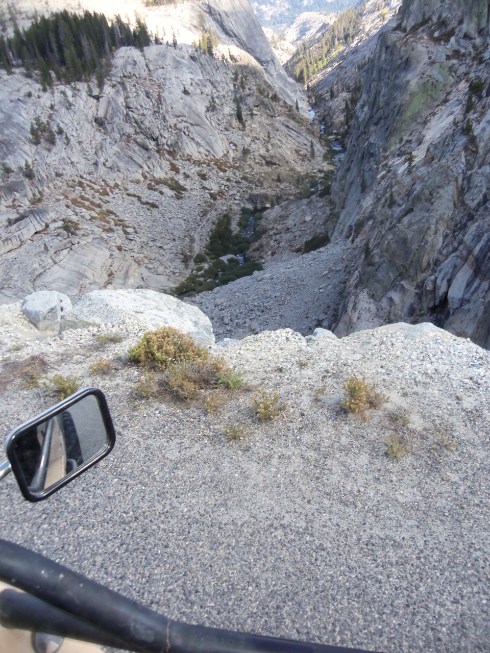 View from a vehicle looking down into a rocky mountain valley with a stream at the bottom. A side mirror is visible