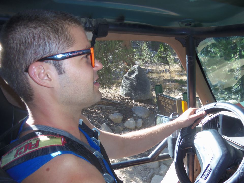 Man wearing sunglasses driving a roll-caged vehicle on a dirt trail