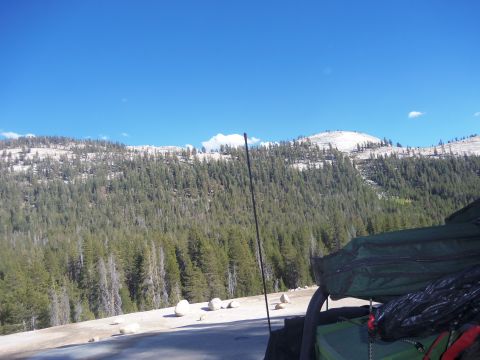 1968 Ford Bronco - Mountain landscape with pine trees and snow-capped peaks under a blue sky. Part of a vehicle with camping gear is visible.