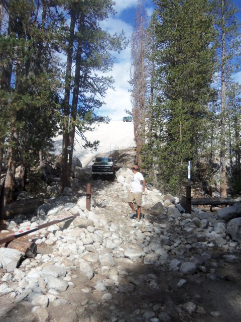 1968 Ford Bronco - Two SUVs navigate a challenging, rocky mountain trail surrounded by pine trees under a bright, cloudy sky.