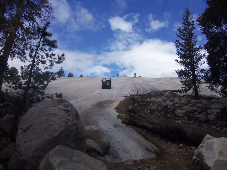 Jeep-like vehicle driving up a snow-covered slope with trees and rocks around