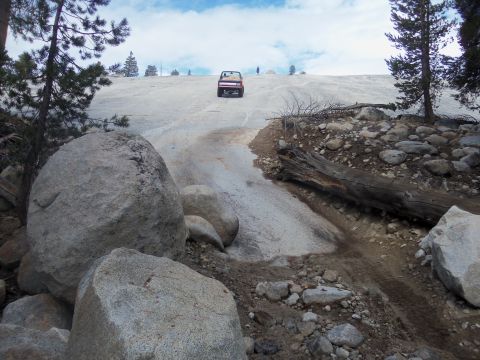 1968 Ford Bronco - A red SUV navigates a steep, snow-covered incline surrounded by rocky terrain and pine trees.