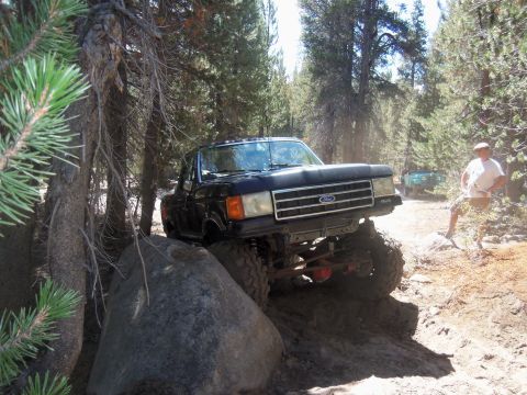 1968 Ford Bronco - A lifted black Ford truck tackles a challenging off-road course, with a second vehicle visible in the distance.
