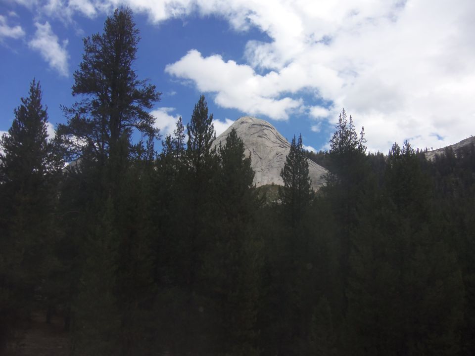 Mountain peak behind dark pine trees under a blue sky with white clouds. Supermotors.net logo in corner