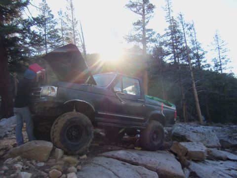 1968 Ford Bronco - A person is attending to the engine of a lifted Ford truck off-road, with the sun shining through the trees.