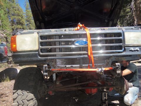 1968 Ford Bronco - A person works on the front suspension of a Ford truck, with the hood open and straps holding the grille.