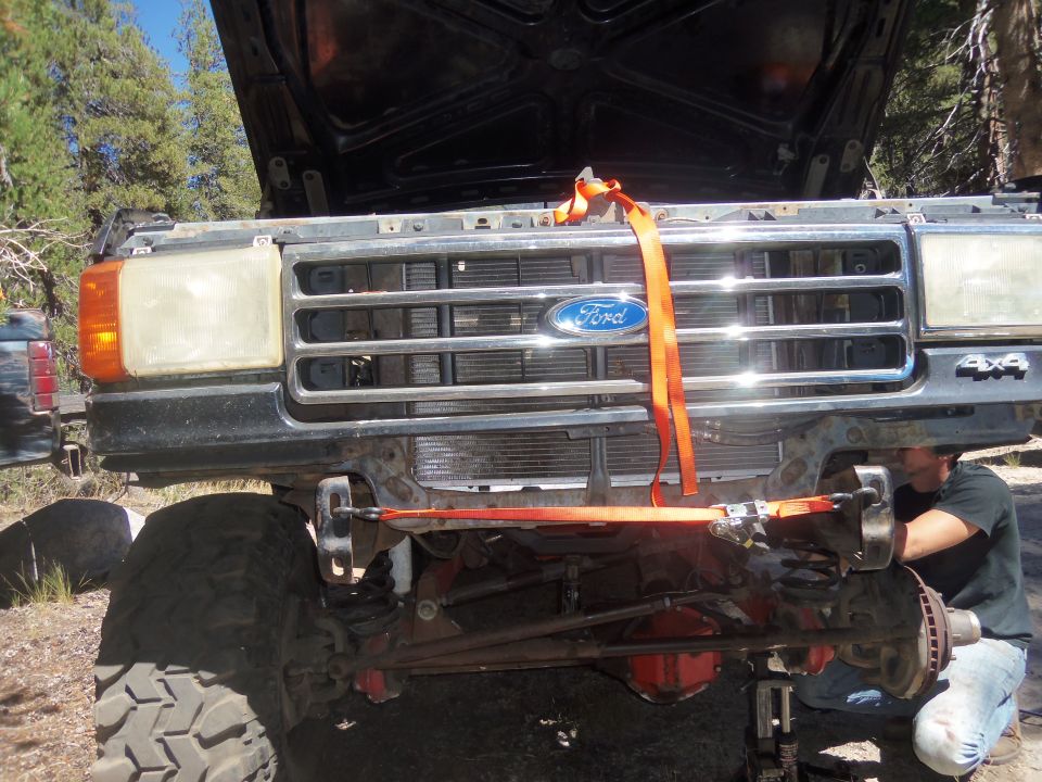 Ford truck front end with hood up, orange straps on grille, 4x4 badge visible. Person working on suspension