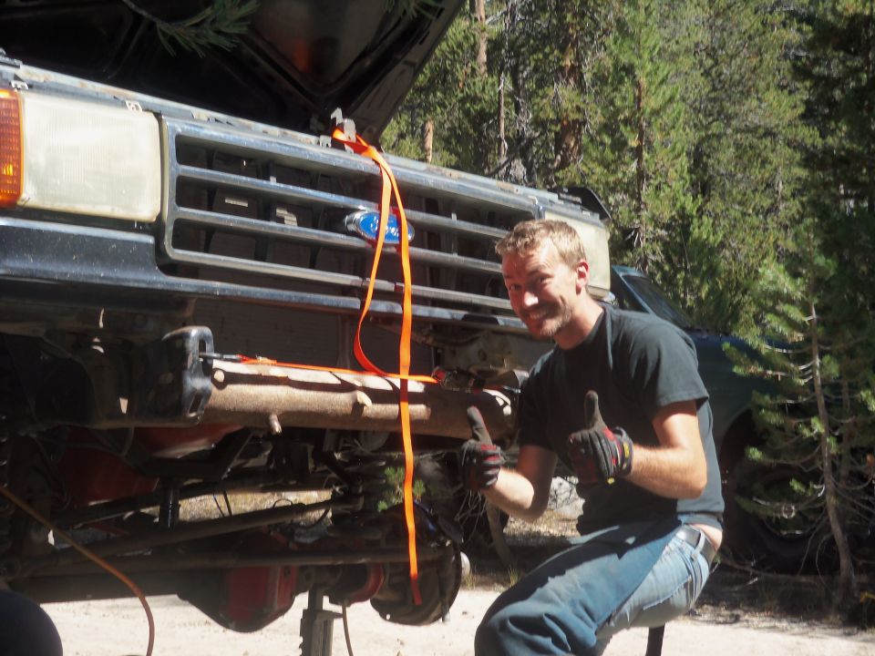 Man gives thumbs up while working on front suspension of a truck; another vehicle is visible in background